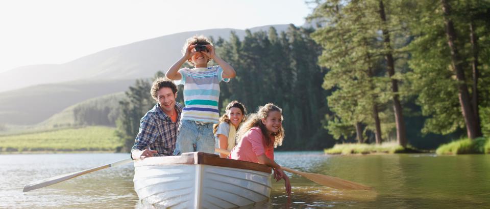 Four people sit in a rowboat on a calm lake; a child stands at the front looking through binoculars, while three adults sit behind, surrounded by trees and hills.