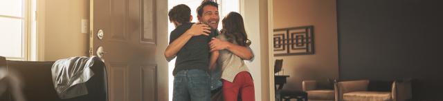 A man stands in the doorway hugging two children in a sunlit living room.